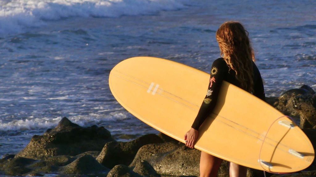 Woman holding a surfboard on the beach at sunset, looking at the ocean — surf school Tenerife