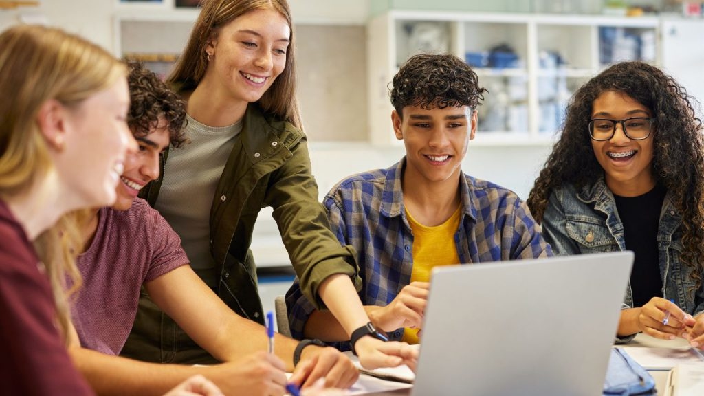 Group of teenagers studying together with a teacher, smiling and practising english collaboratively on a laptop in a supportive classroom environment.