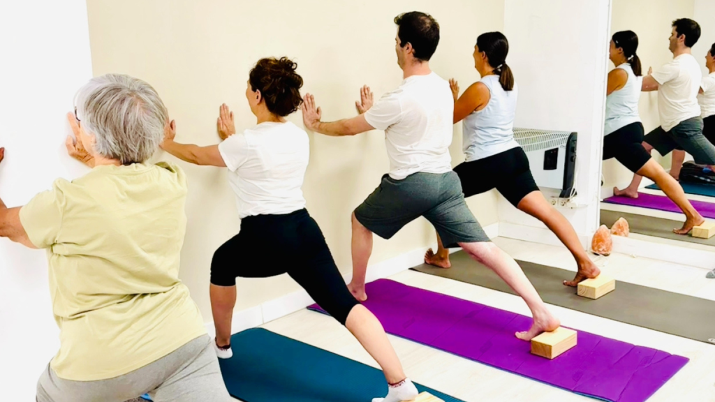 Clase grupal de yoga iyengar con alumnos de distintas edades practicando posturas de pie con apoyo en la pared y bloques para mejorar alineación y estabilidad corporal